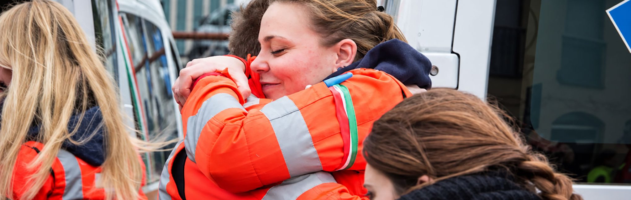 Volontari ANPAS in divisa arancione si abbracciano accanto a un’ambulanza